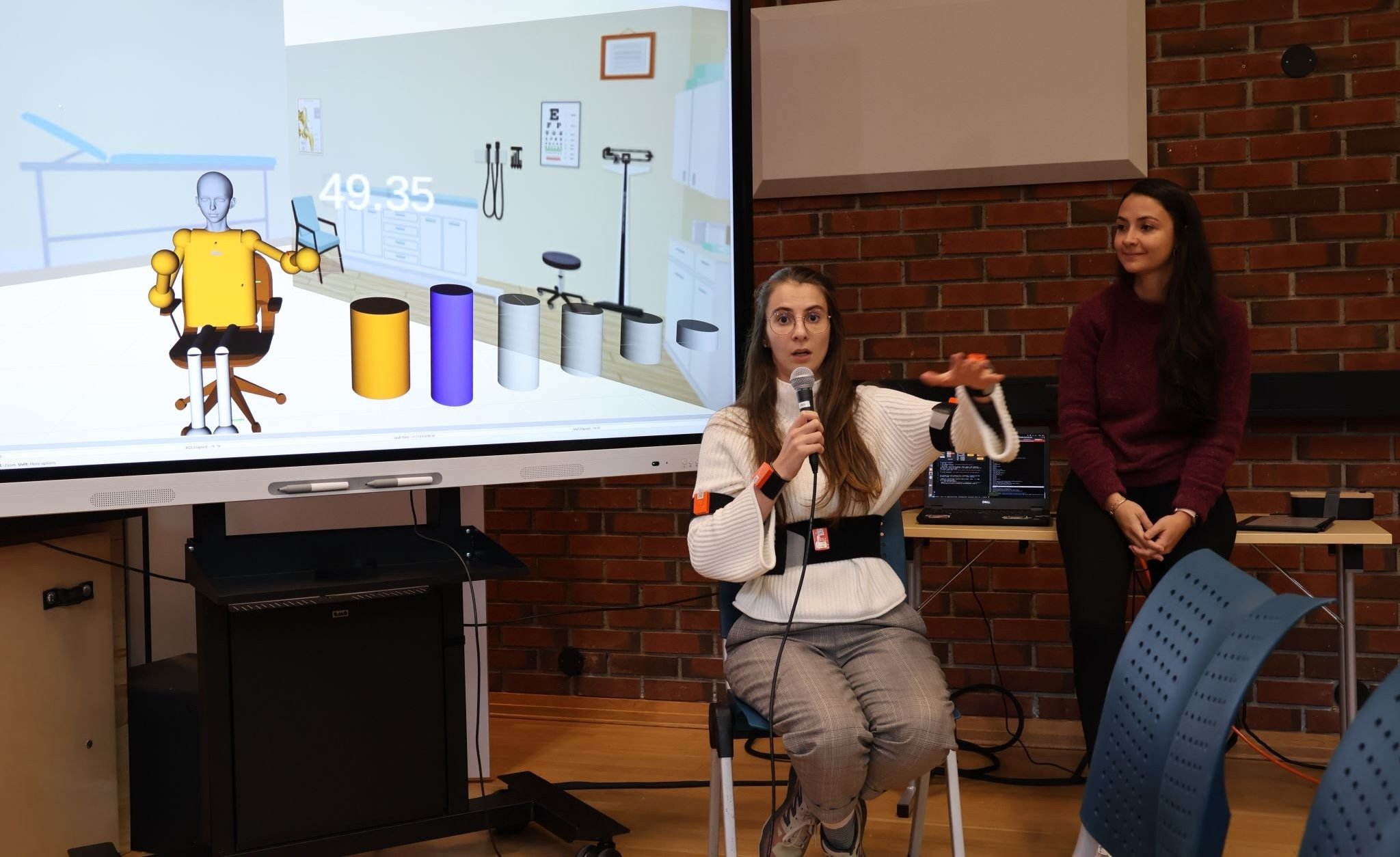 Raquel sitter på en stol og har festa på seg sensorer. Hun løfter den ene armen. På en skjerm i bakgrunnen ser vi en digital figur som løfet armen på samme måten. Til høyre i bildet står Gizem og ser mot Raquel.