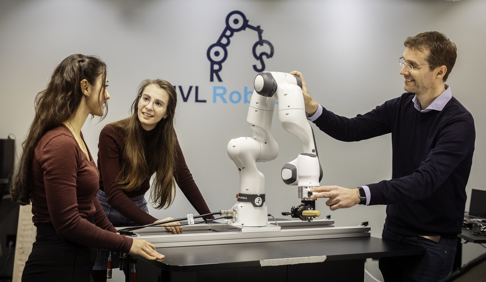 Tre personer står ved et bord og ser mot en robotarm som står på bordet. 