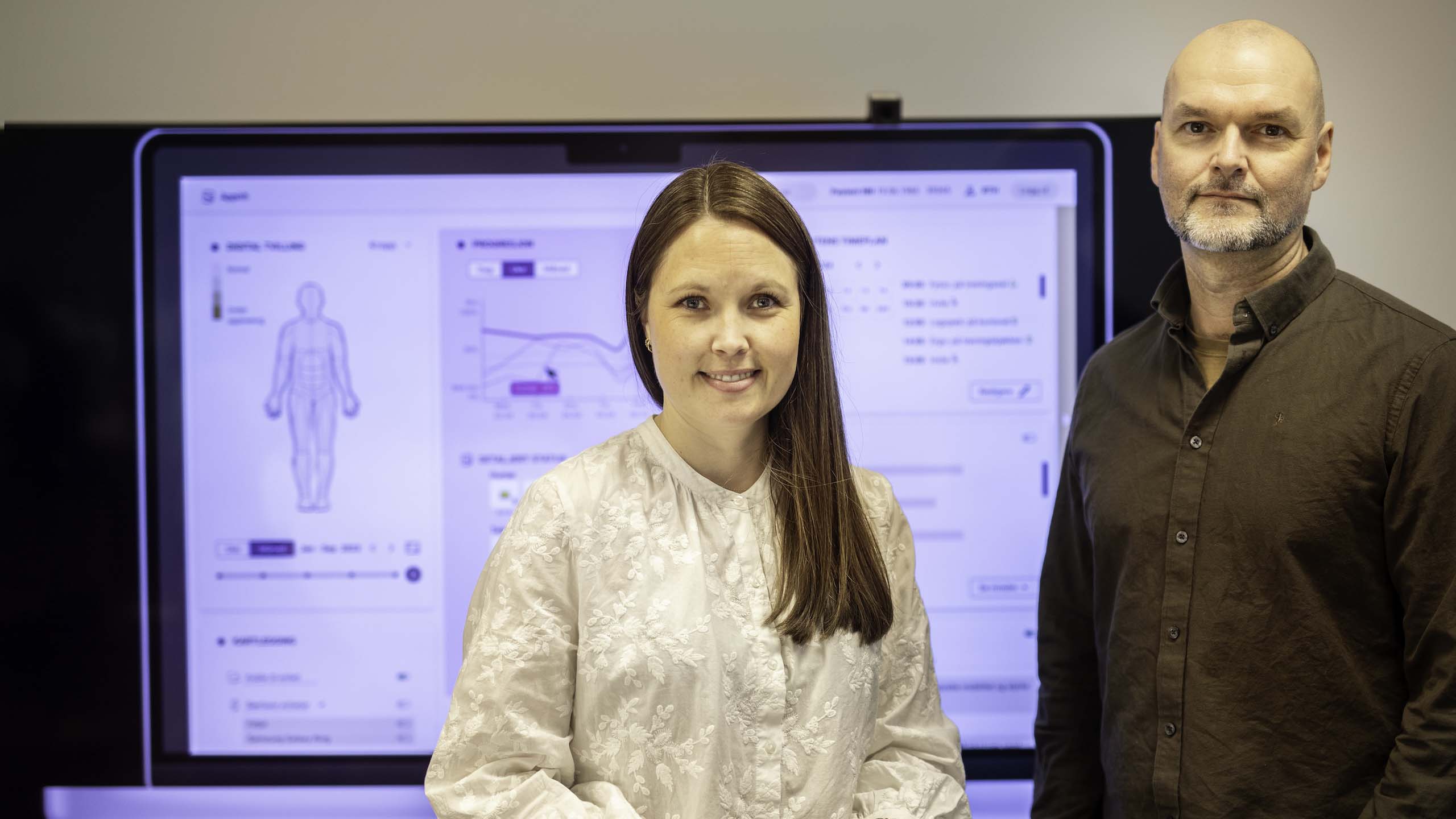 Kvinne og mann står og ser mot kamera. I bakgrunnen ein skjerm som viser ein modell av ein tenkt digital tvilling - ein menneskefigur som skal spegle tilstanden og utviklinga til pasienten som blir målt.