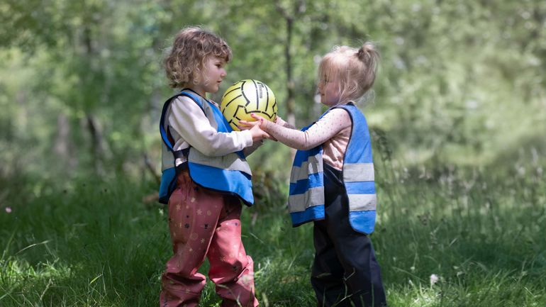 To barn i regnbukser og vest står mot hverandre. En av dem holder en ball. 