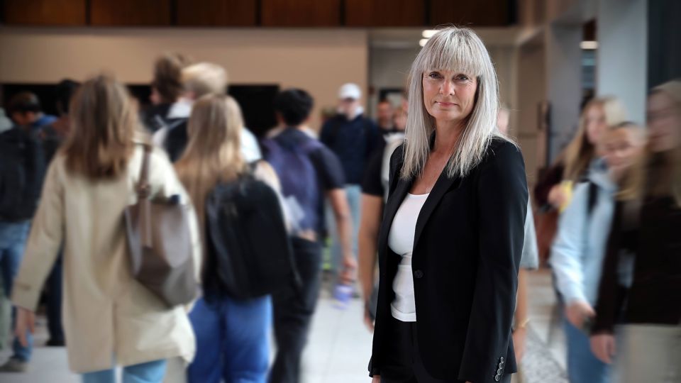 Ei kvinne (Milfrid Tonheim) står og ser inn i kamera og smiler. Ho er i eit inneareal på høgskulen og plassen er travel, med fleire studentar som går forbi med sekkar og veskar.