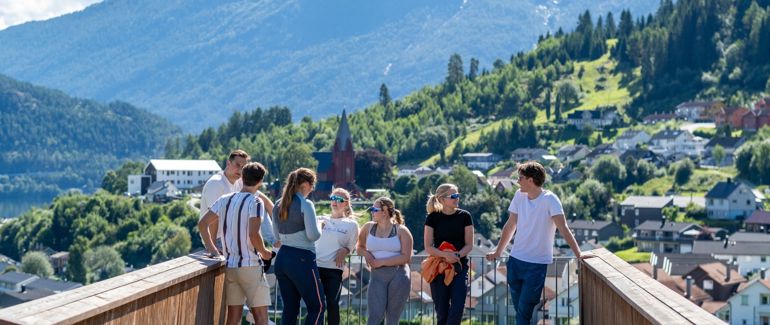 Syv studenter står på et utkikkspunkt på Campus Sogndal, hvor utsikten er store fjell, frodig grønt landskap, og noen hus og en kirke.