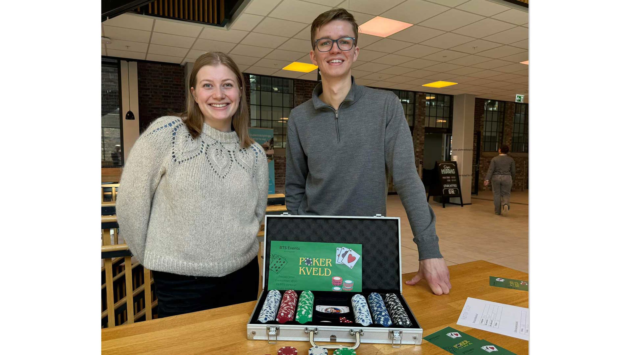 To studentar smiler til kamera. På bordet framfor dei står det ein koffert med poker-sjetongar, samt ein poster for eit pokerkveld-arrangement.