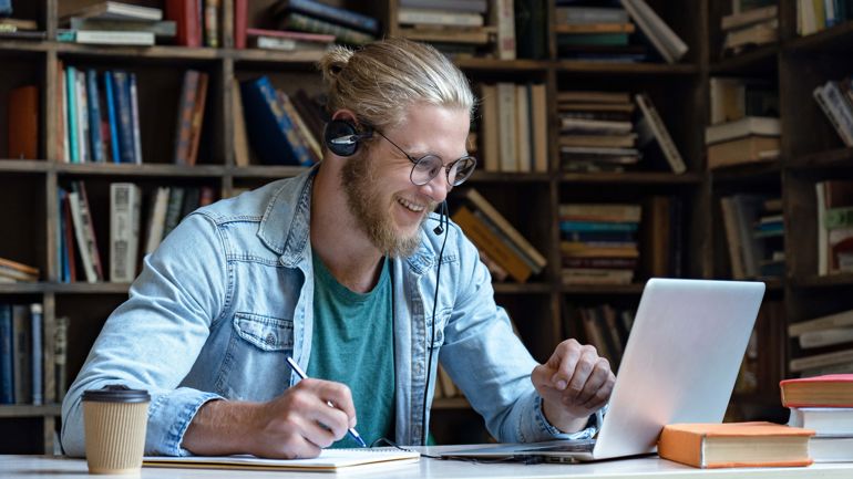 Mann sitter foran bærbar pc. Han har hodetelefoner på hodet, og han noterer i en notatbok. Bak ham er det mange bokhyller. 
