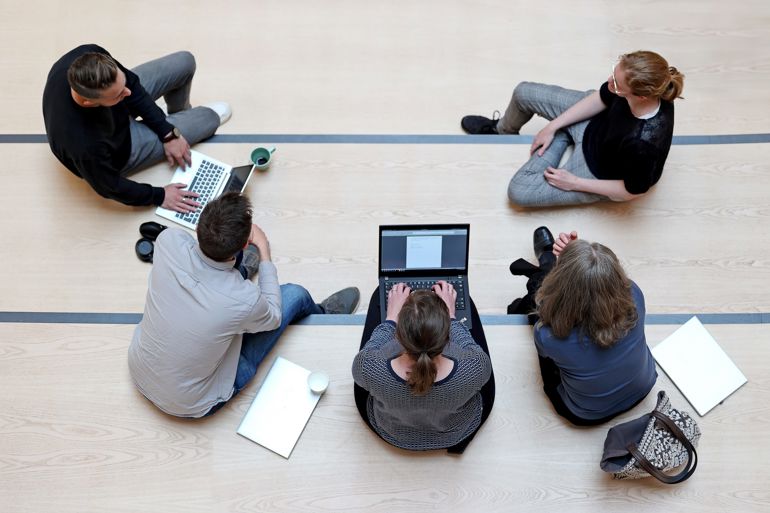 Et fugleperspektiv av fem personer i en halvsirkel som diskuterer og samarbeider med PC-er og notater på gulvet