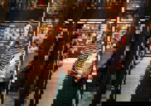 Professor Edit Bugge står i døra til biblioteket på campus Bergen. Ho smiler. Edit har raudleg hår og er kledd i ein genser med svarte og oransje striper og eit grønt skjørt. Ho held eit utval bøker av ulik storleik.