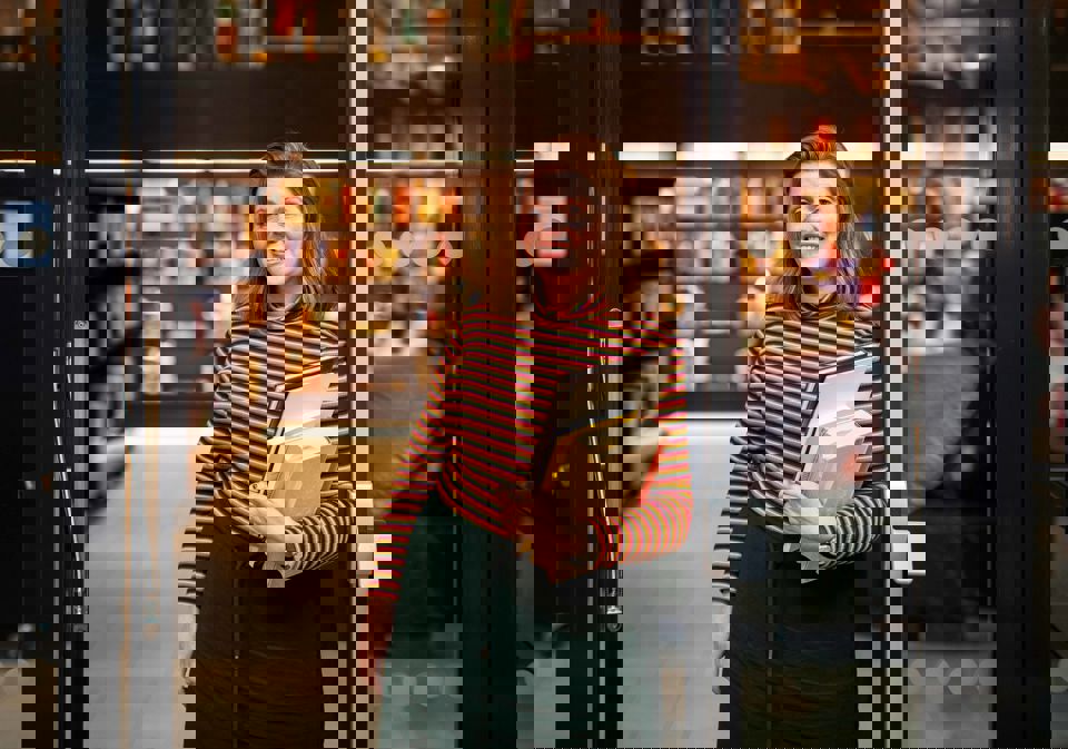 Professor Edit Bugge står i døra til biblioteket på campus Bergen. Ho smiler. Edit har raudleg hår og er kledd i ein genser med svarte og oransje striper og eit grønt skjørt. Ho held eit utval bøker av ulik storleik.