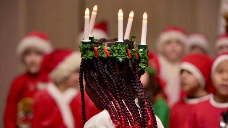 Jente med melaninrik hud og lange fletter har Lucia-krone på hodet. Barn i bakgrunnen med nissedrakter.
