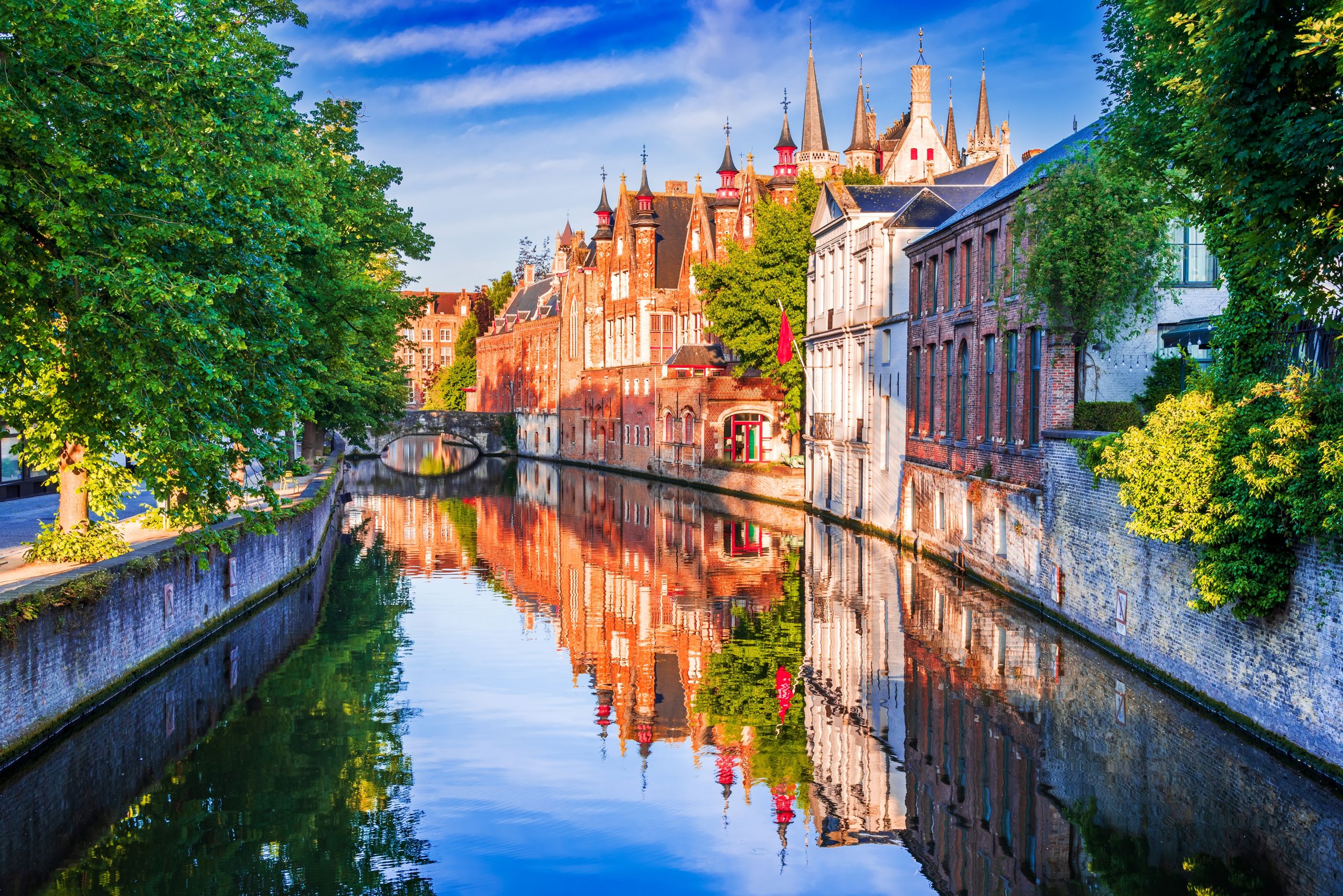 Bilde av Brugge i Belgia, en kanal gjennom byen, med eldre bebyggelse på høyre side, trær på venstre side og en bro foran i horisonten.