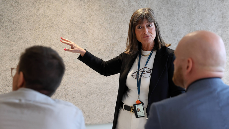 Kvinne står foran to menn i et klasserom og peker mot tavle