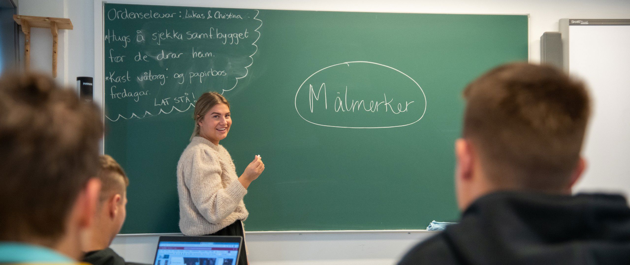 Lærer som står til venstre for tavlen foran en skoleklasse med kritt i hånden.