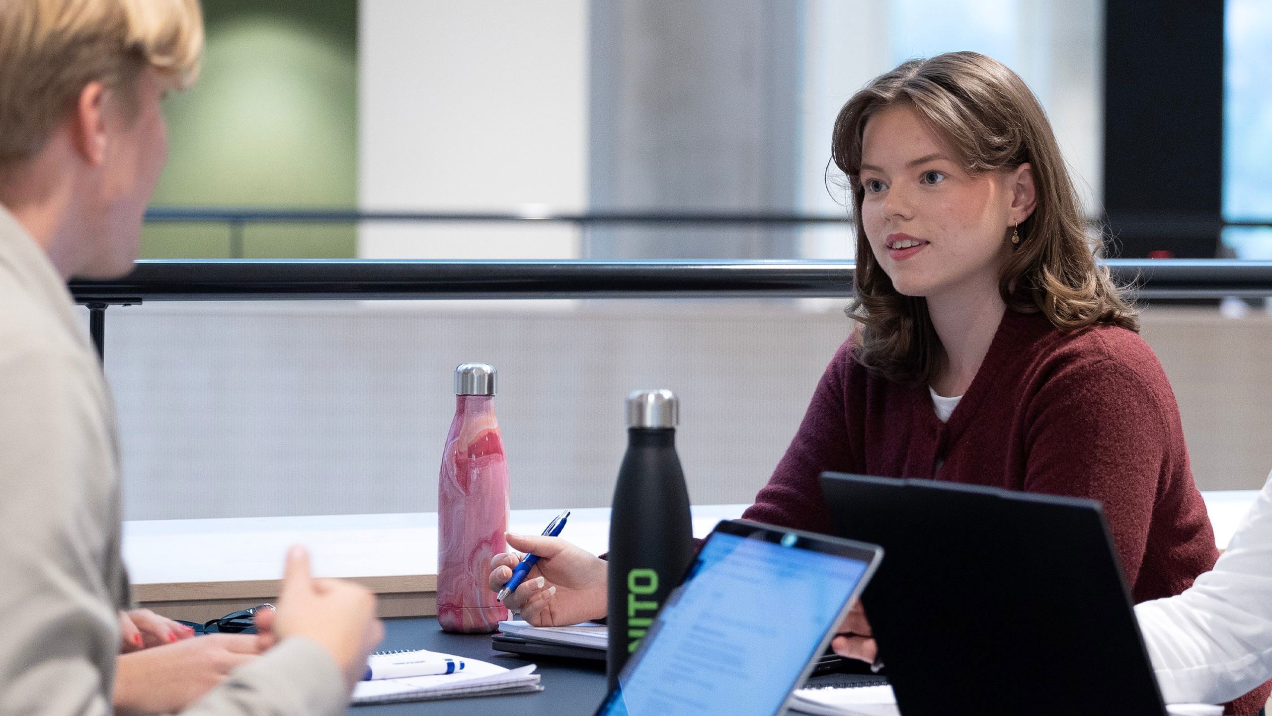 To studenter sitter ved et bord, en jente med brunt skulderkort hår kikker bort på andre siden mot en mannlig student. De snakker og skriver både på pc og i blokk. De har også vannflasker på bordet i aleminium.