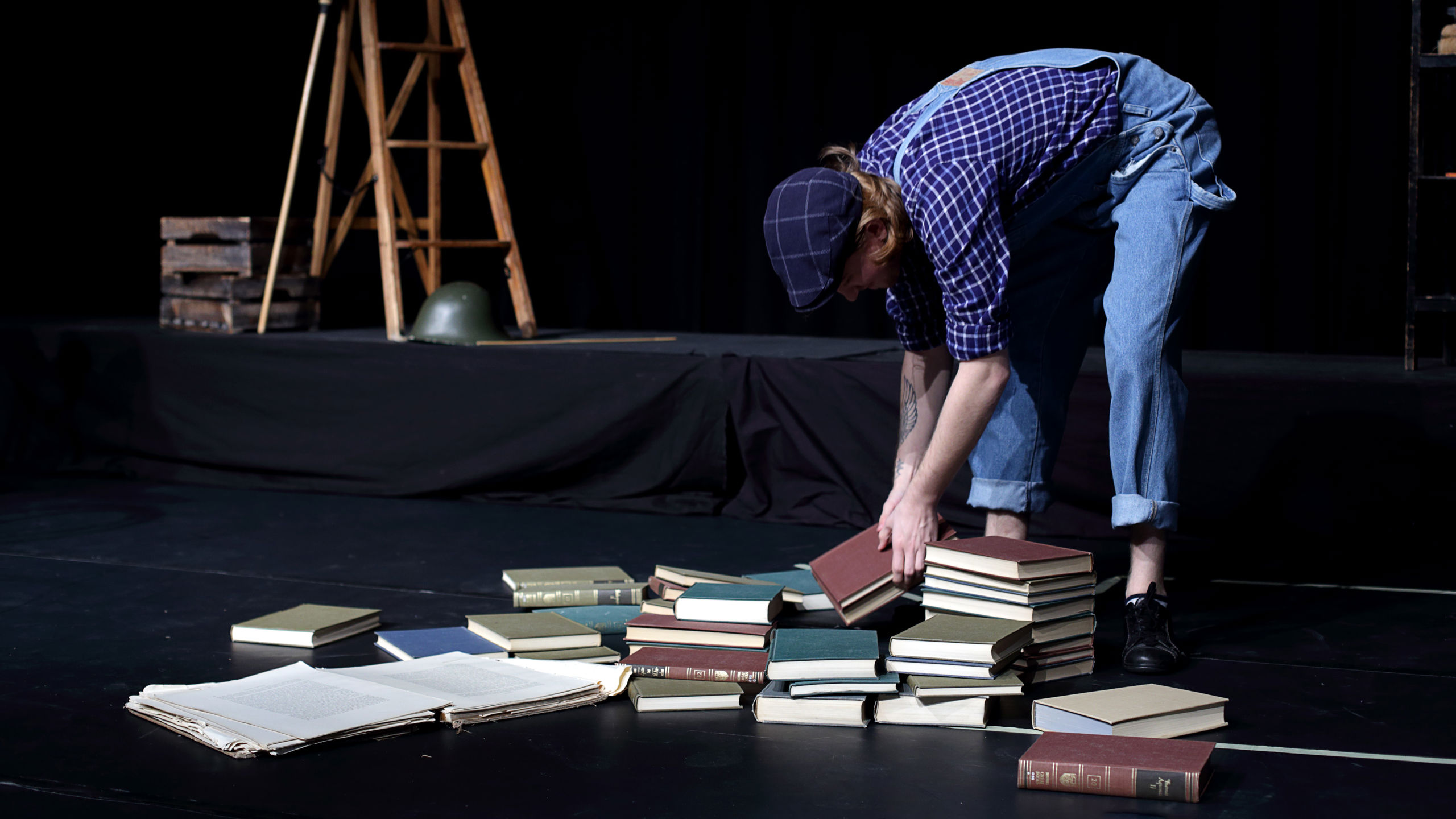 Eit bilde frå ein scene. Ein person med raudblonde krøller, kledd i snikkarbukser, blå- og kvitruta skjorte og ein blå sixpence, står bøygd over in haug med bøker som ligg på scena.