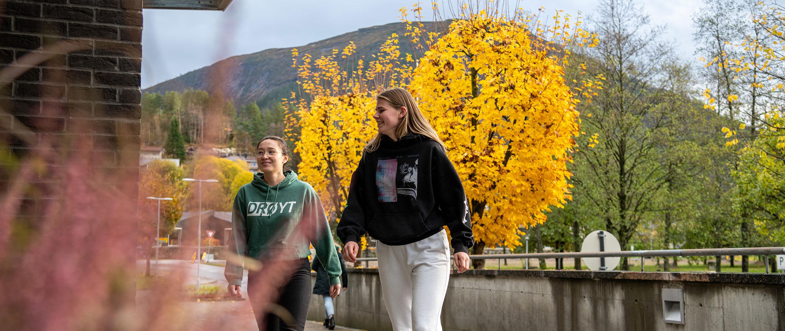 To jenter utanfor campus med gule haustblad i bakgrunnen. 