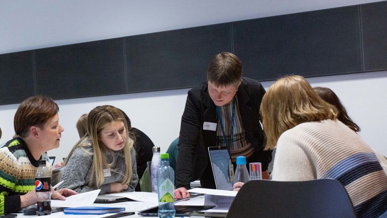 Gruppe mennesker jobber sammen rundt et arbeidsbord. En dame står over de og instruerer. 