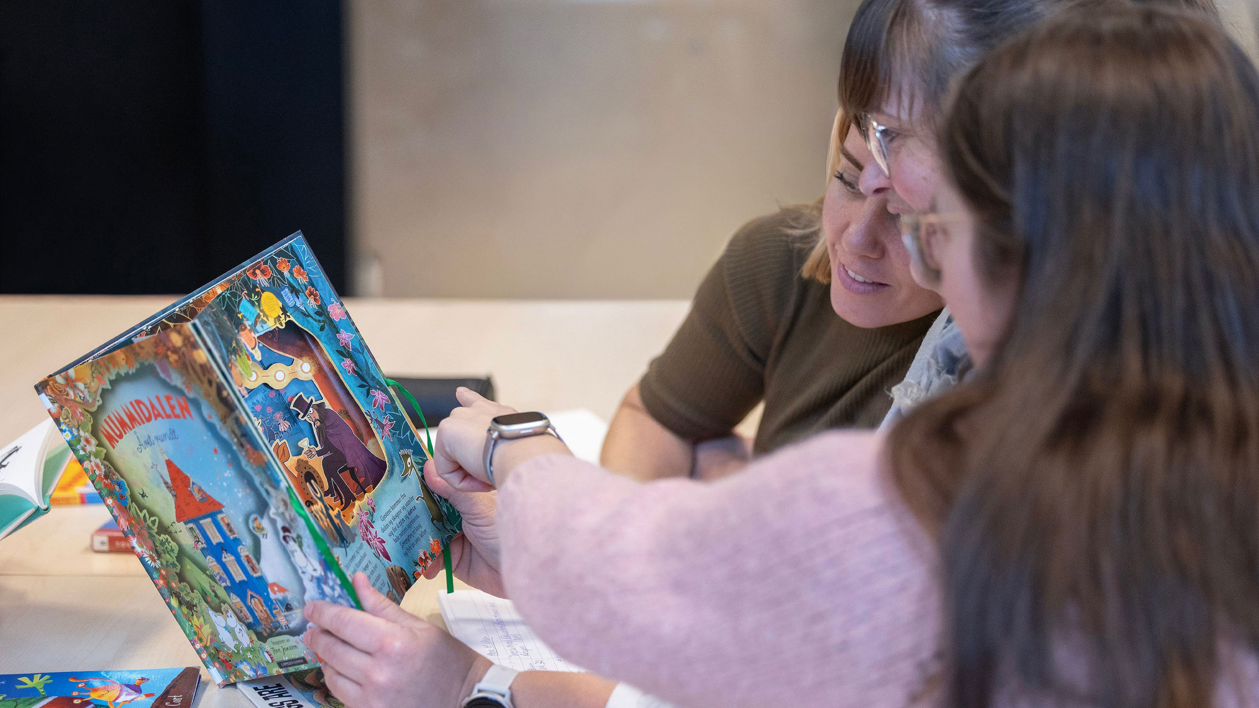 Tre studentar, plassert til høgre i bildet, sit og blar i ei fargerik Mummidalen-bok. Ei av dei peiker på noko i boka, og dei to andre ser engasjert på boka.