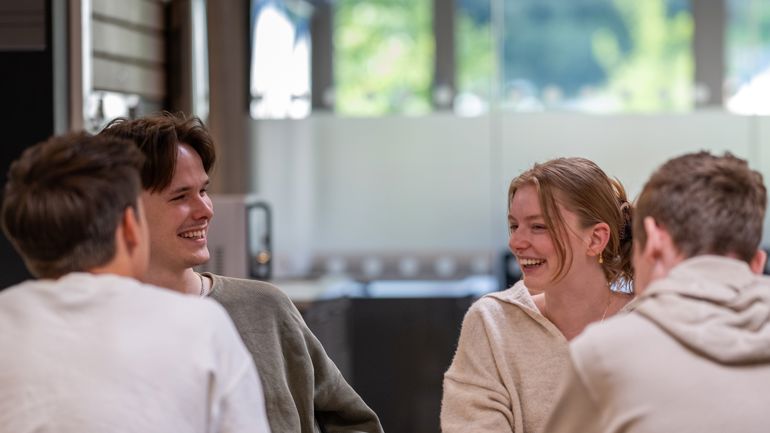 Fire studenter sitter rundt et bord. To av guttene er med ryggen til kameraet, og en gutt og en jente er i fokus. De smiler bredt mot hverandre.