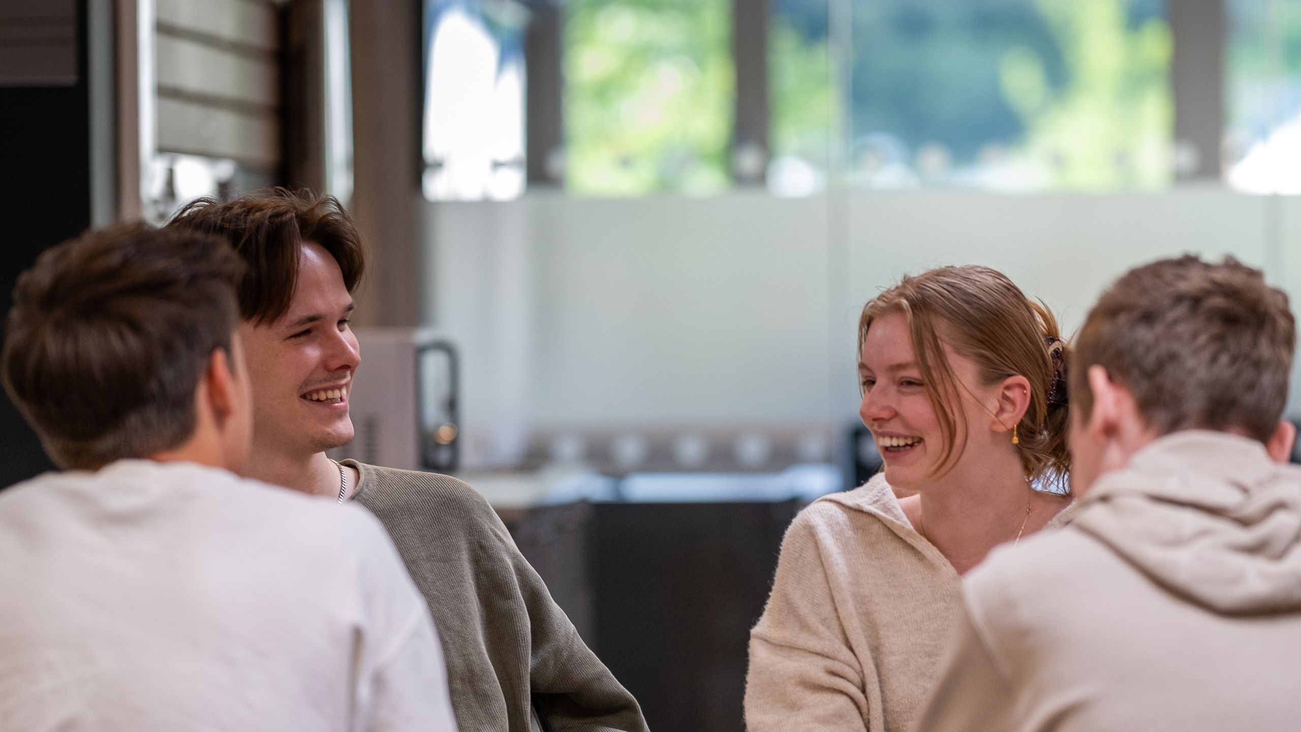 Fire studenter sitter rundt et bord. To av guttene er med ryggen til kameraet, og en gutt og en jente er i fokus. De smiler bredt mot hverandre.