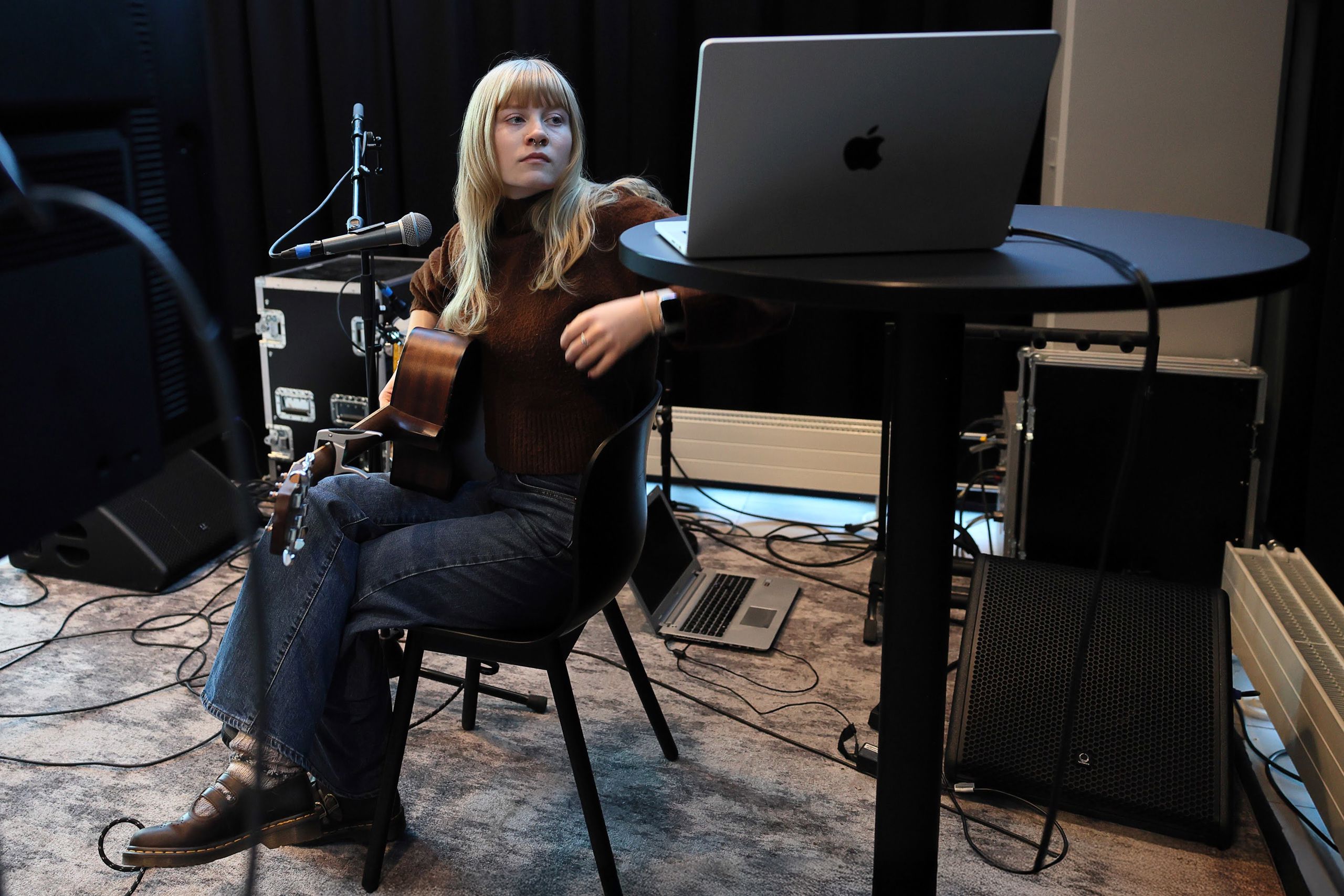 Ein person med langt, blondt hår, pannelugg og septumpiercing sit på ein stol og ser på ein PC som står på eit høgt bord. Personen har ein gitar i fanget og ein mikrofon føre seg.