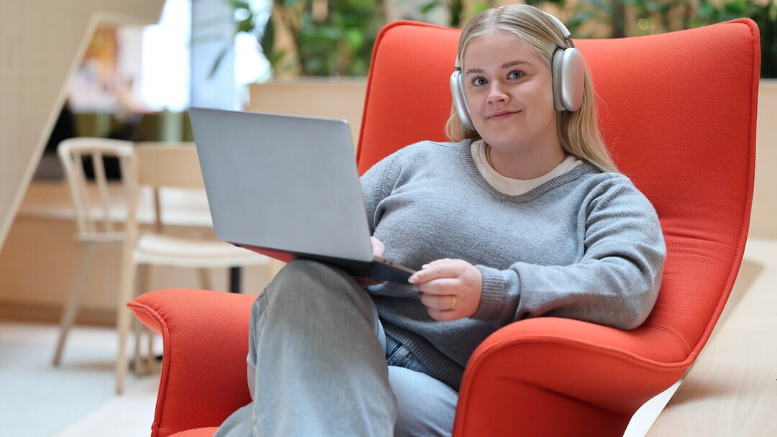 Ung kvinne sitter i lenestol med pc i fanget og headset på ørene. 