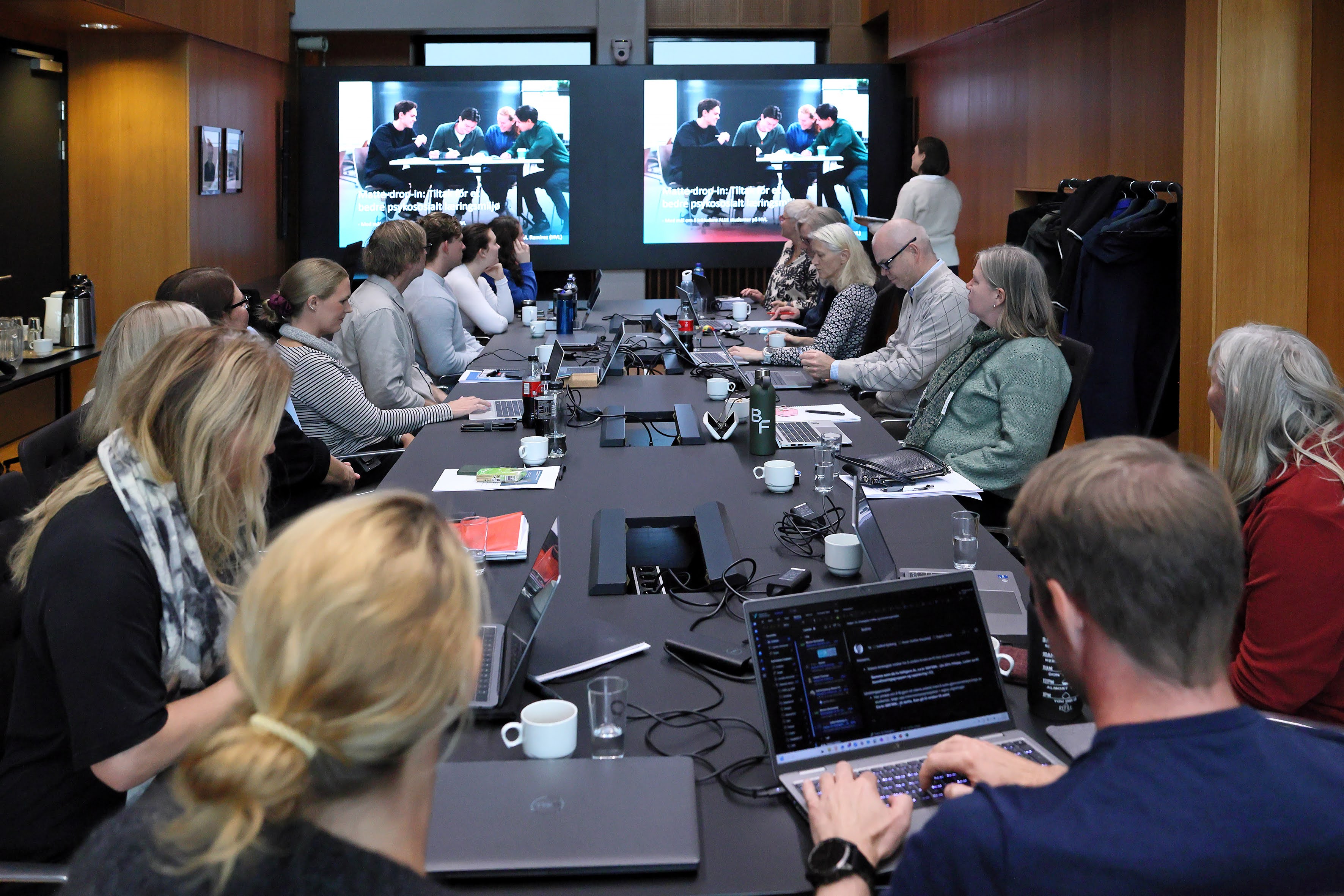Ei gruppe menneske sit rundt bordet i eit møterom. Dei følger med på ein presentasjon som går på to skjermar fremst i rommet. På skjermane ser ein ein gjeng unge menneske som sit rundt eit bord og snakkar.