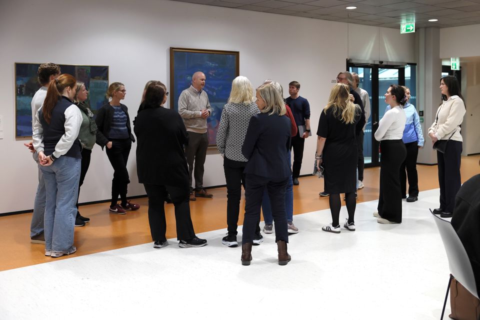 Ei gruppe menneske står samla rundt ein kar, Endre Laastad, som snakkar med dei frammøtte. Dei står i eit rom med brunoransje og kvitt golv, kvite veggar og kunst på veggane.