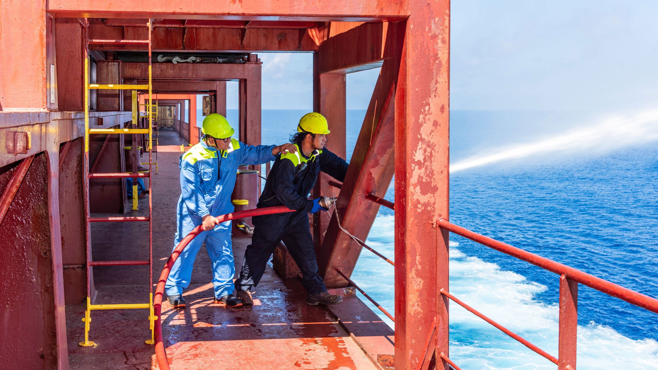 To menn med hjelm og verneutstyr på et skip. De holder i en brannslange. 