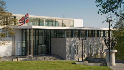Eit moderne, kvitt bygg med store vindauge. Framfor bygget står ei flaggstong med det norske flagget heist høgt.