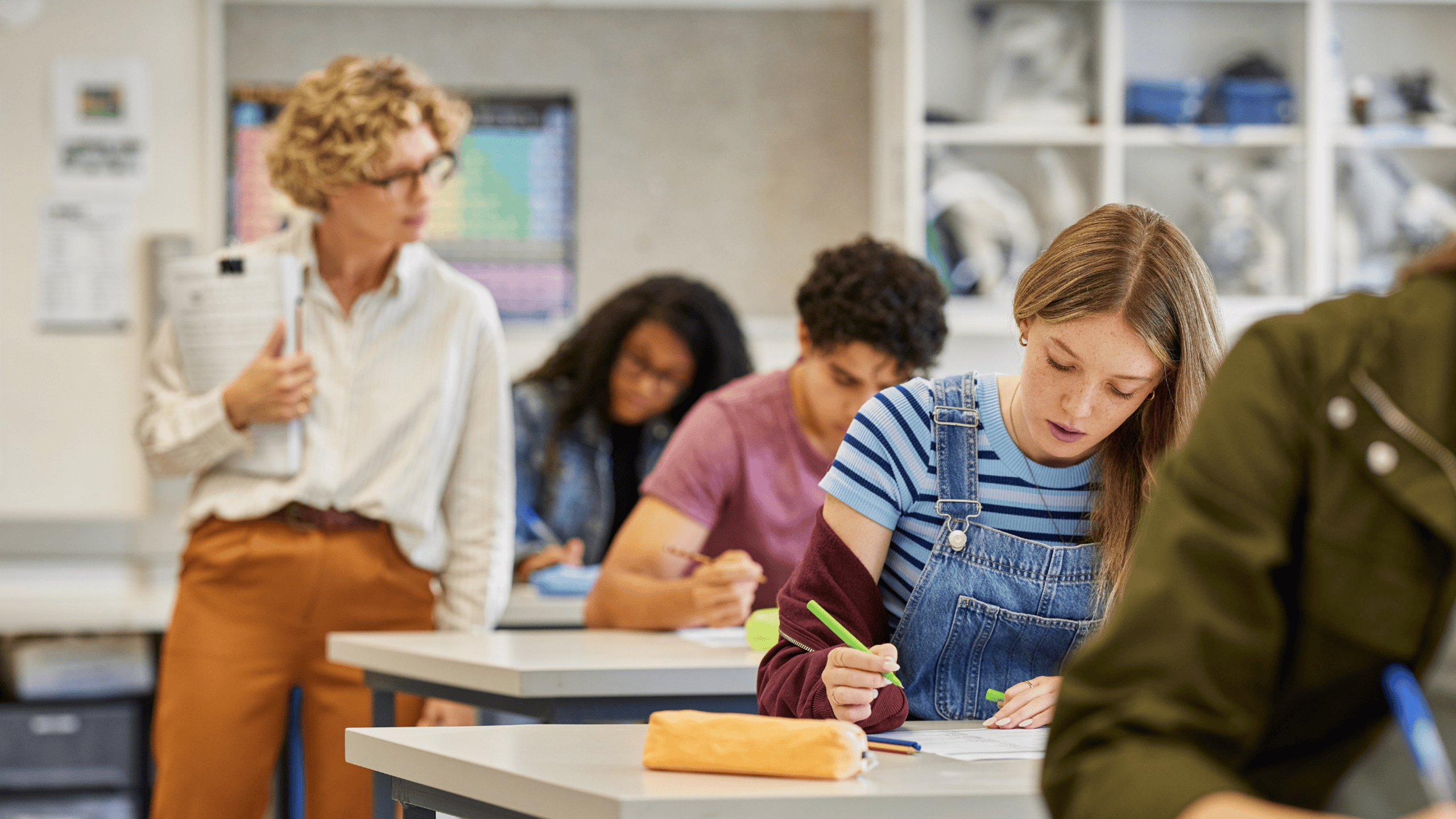 Moden lærer overvåker elevene mens de skriver eksamen i et klasserom på ungdomsskolen. Gutter og jenter skriver på eksamensark under lærertilsyn.