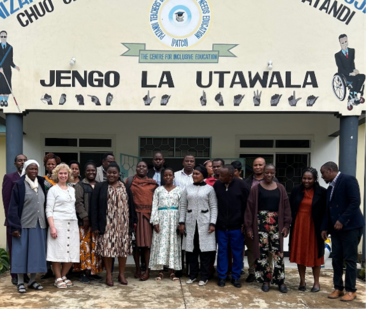 Ein gruppe personar står utanfor ein bygning. På veggen over dei står det: Jengo la Utawala. 