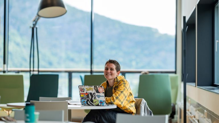 Jente som sitter på biblioteket i Sogndal, store vinduer i bakgrunnen som viser grønn natur. 