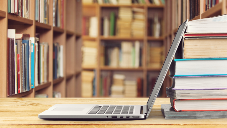 Stabel med bøker foran en pc, i bakgrunnen er det bokhyller med masse bøker