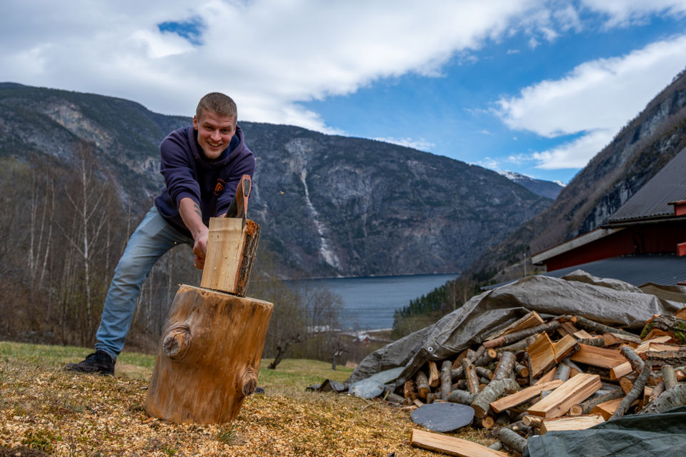 Mann som hoggar ved, med fjord og fjell i bakgrunnen.