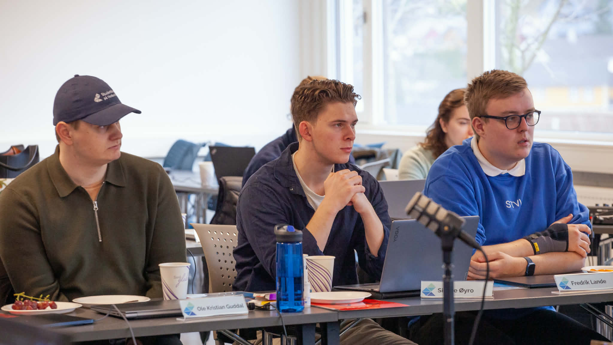 Tre menn sit ved eit bord. Mannen til venstre har caps.