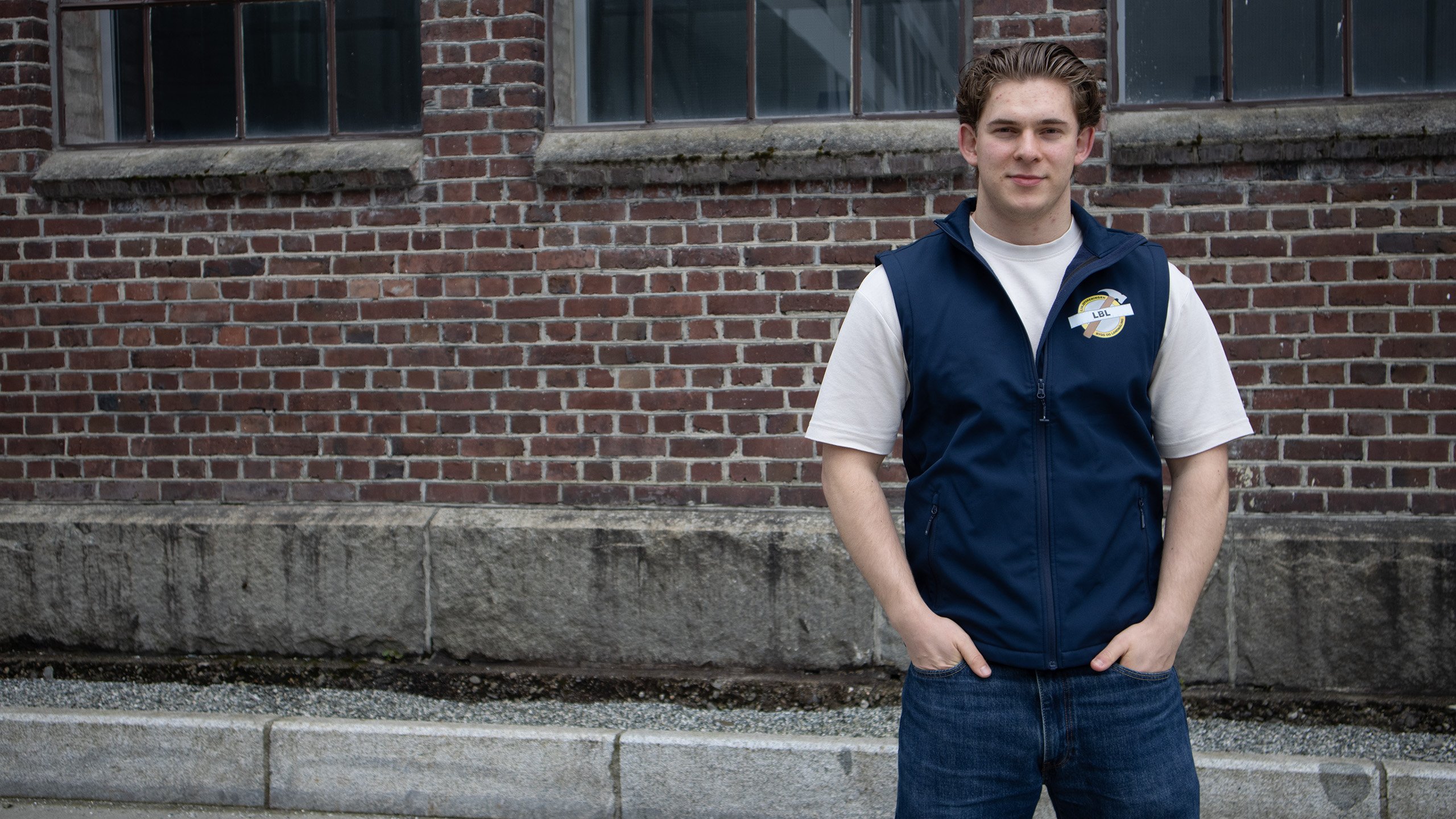 Mann i T-skjorte og vest står foran murvegg.