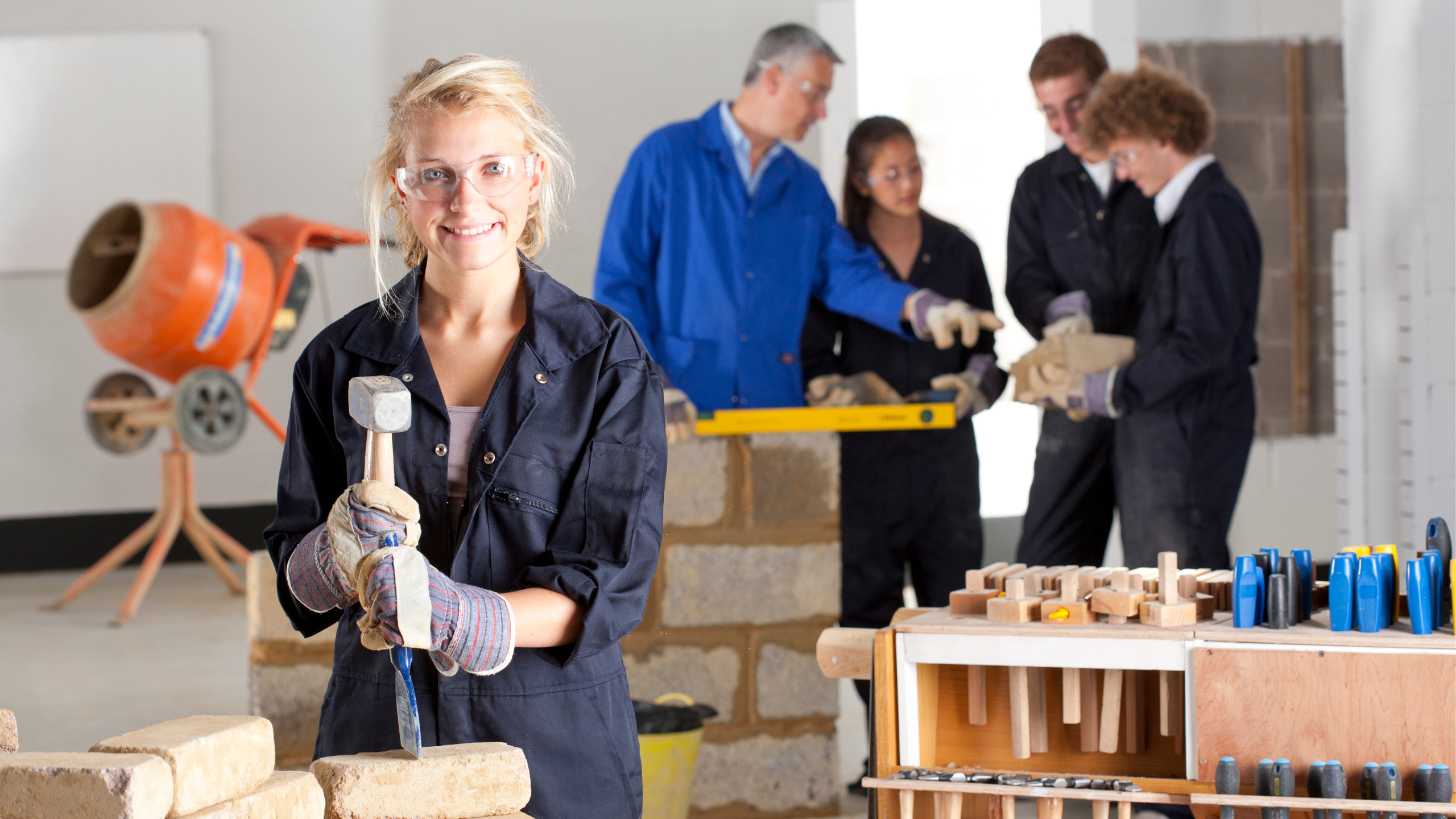 Girl in work clothes and safety glasses with tools with people in the background