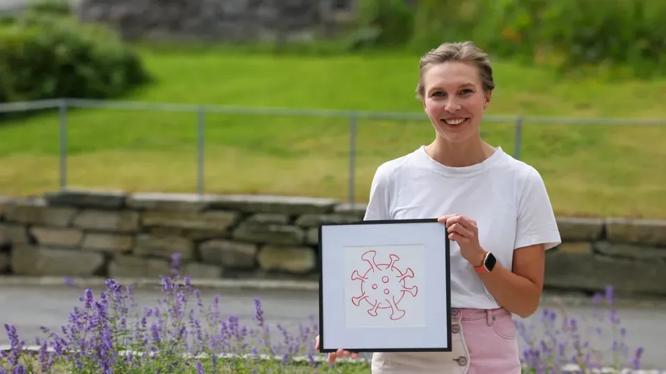 Forskar Kiri Lovise Njøten, blond og kledd i lyse fargar, held opp ein illustrasjon av eit koronavirus.