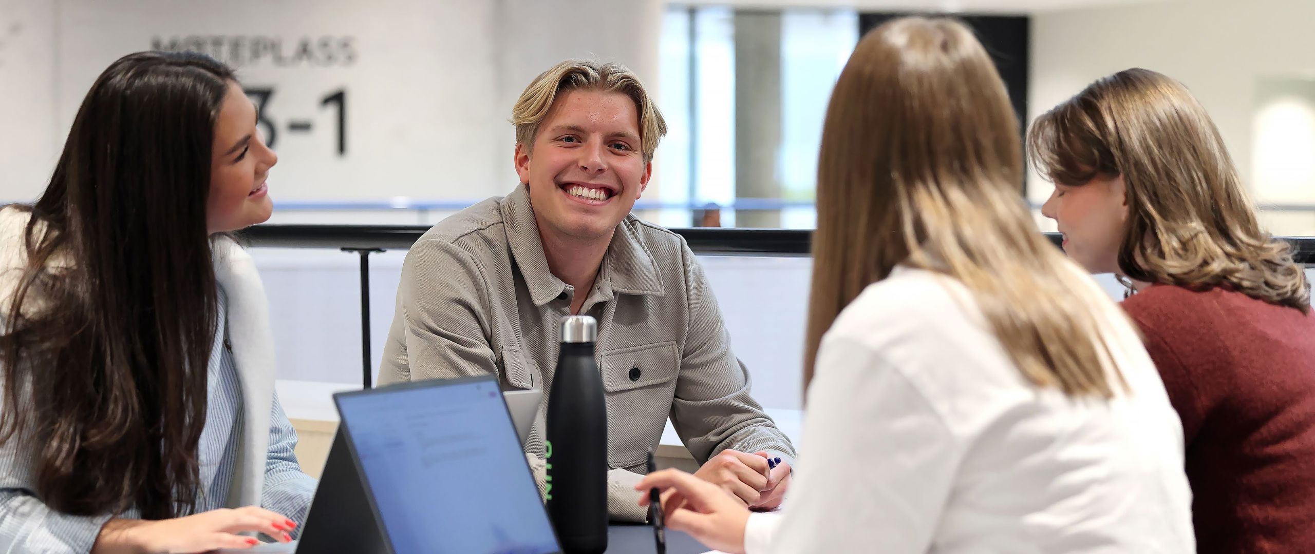 Four students around a table at K1, Campus Bergen. A blond boy looks into the camera and smiles broadly.