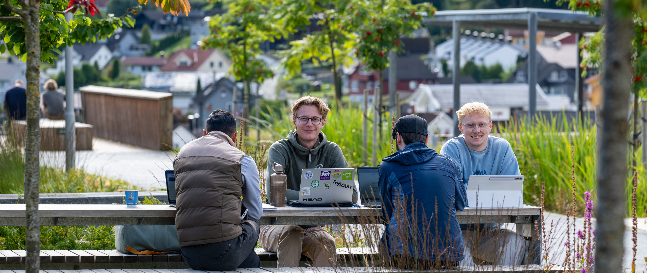 Fire gutter sitter ute på benk med bord på campus Sogndal. To av dem med ryggen i mot, og to med ansiktet til kameraet. Pc-er på bordet med flere klistremerker på fra ulike studentorganisasjoner.