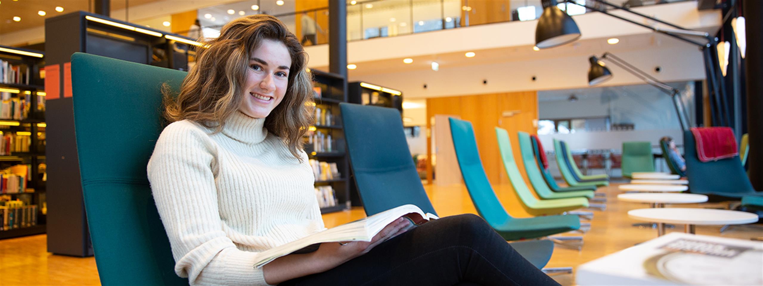 Student sittande i stol på biblioteket med ei bok.