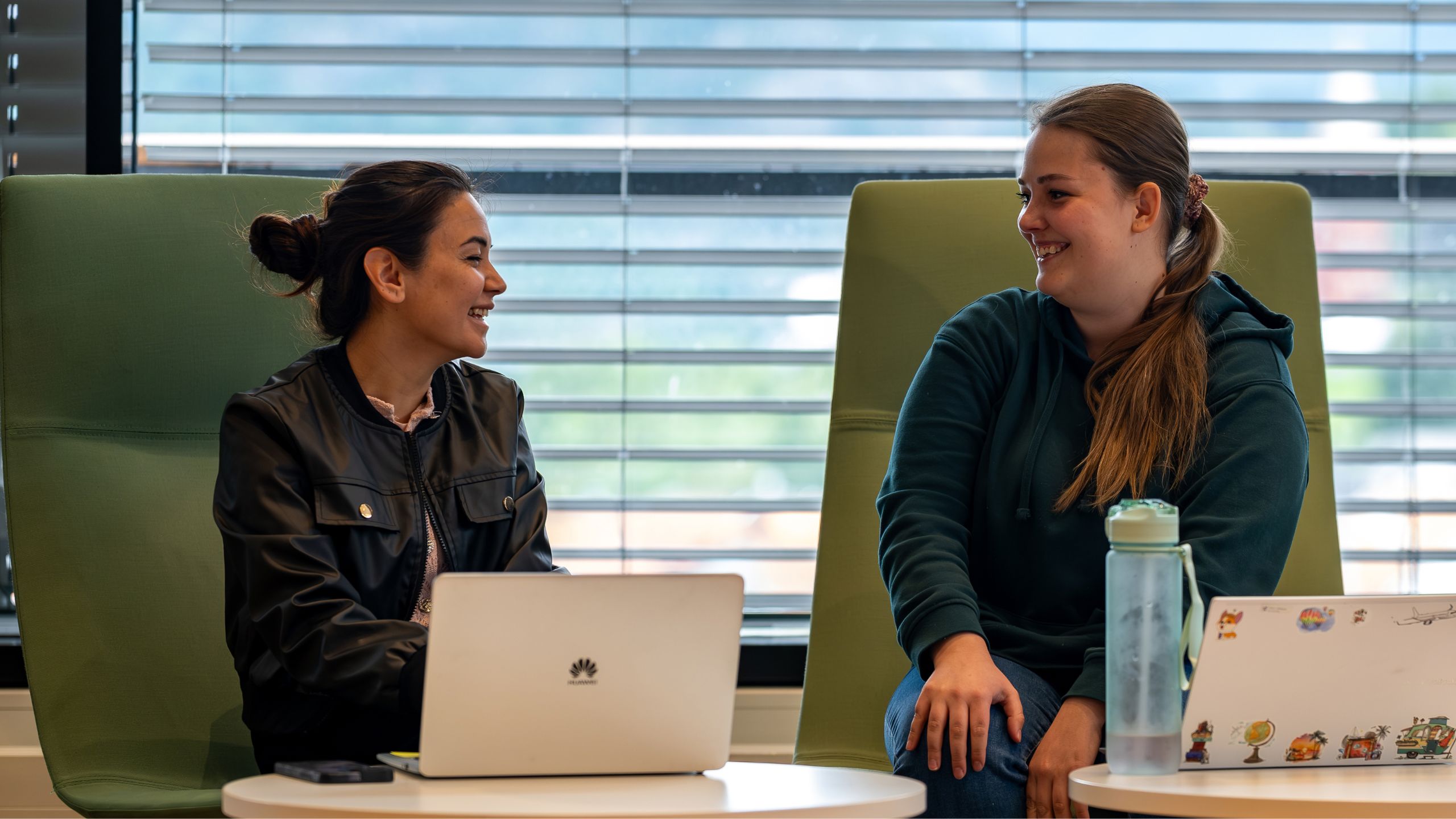 To kvinnelige studenter som sitter i to store stoler på biblioteket i Sogndal. Begge ser på hverandre og smiler. 