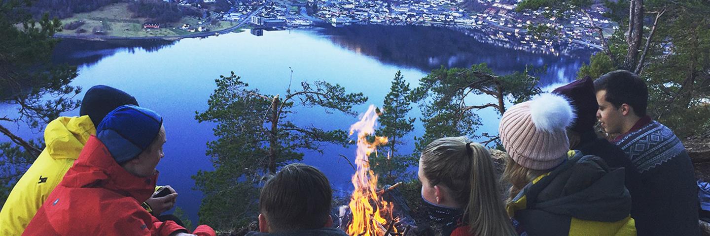 Studentar på båltur i skogen med fjorden og Sogndal i bakgrunnen. 