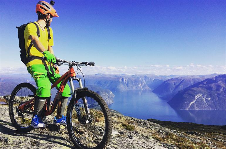 Syklist på fjelltopp med fjord og fjell i bakgrunnen. 