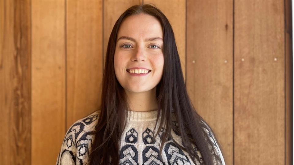 Student står foran en trevegg og smiler til kameraet. 