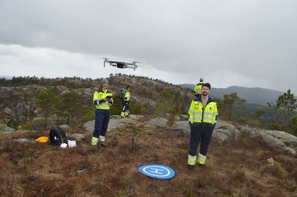 Bildet viser fire mennesker kledd i i arbeidsdress oppe på et fjell. En kvinne styrer en drone i lufta. Hun smiler.