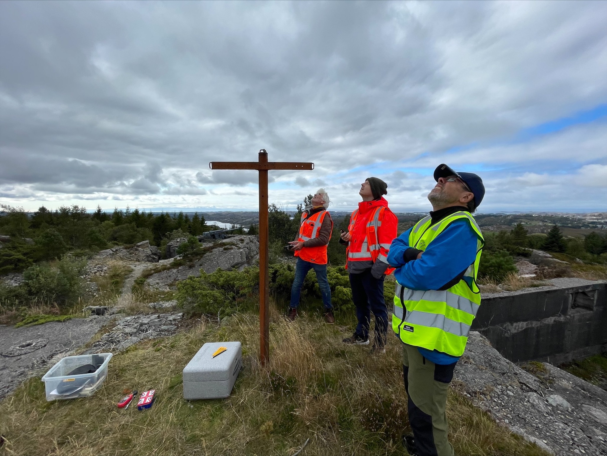 Bildet viser tre menn på toppen av en høyde utendørs. De kikker opp i luften på en drone. 