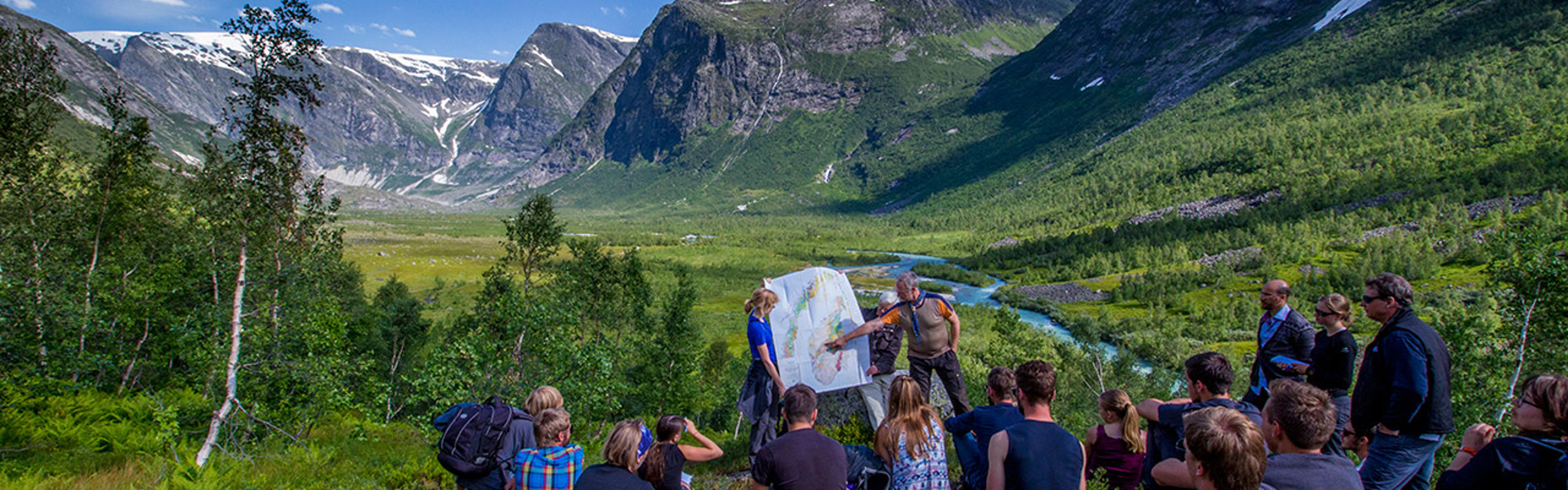 Students on excursion sitting in the gras, surrounded by mountains and blue sky. 