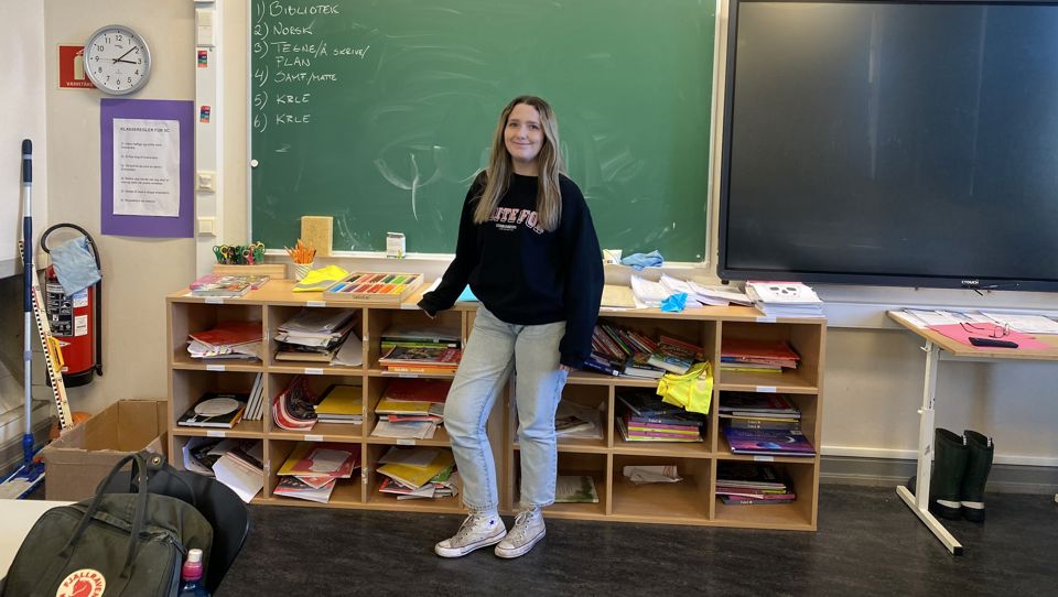 Ei ung kvinne står fremst i eit klasserom, framfor ei tavle, og smilar mot sjåaren.