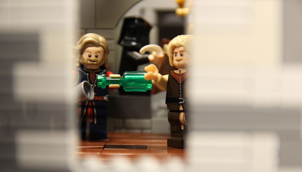 Vi ser inn gjennom eit vindue, der to Lego-figurer står i lag og ser glade ut. Den ene skjenker noko frå ei grøn flaske og ned i eit beger.