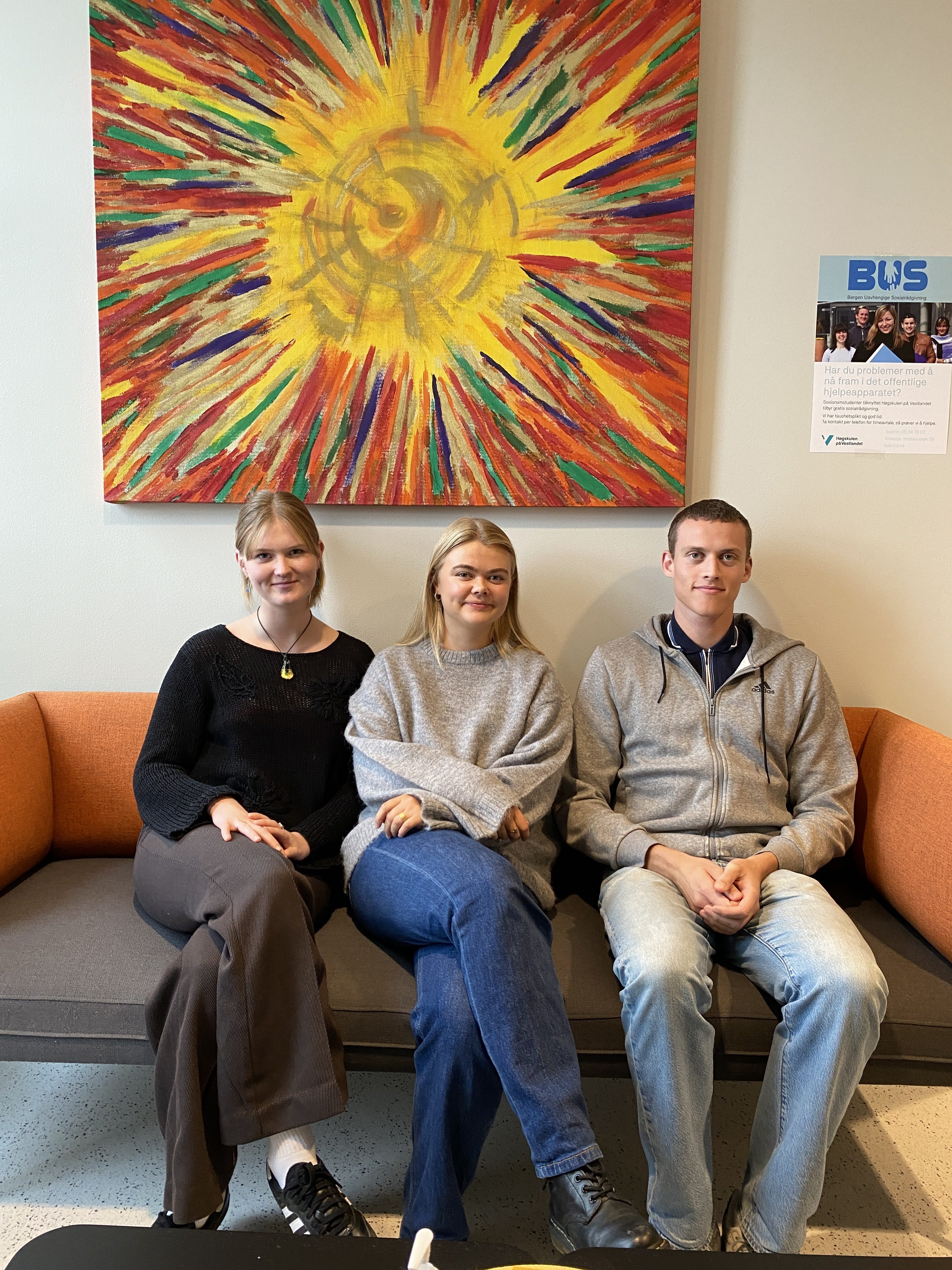 Tre unge mennesker i en sofa, og et stort maleri på veggen bak dem.